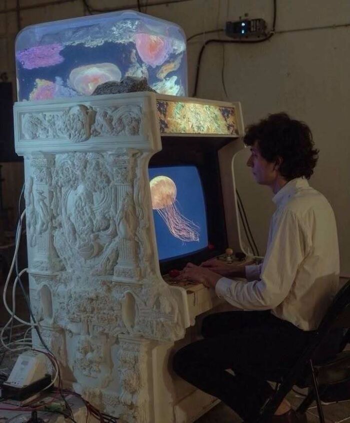 Person playing arcade game with intricate classical sculpture design and jellyfish imagery, blending trash and culture.