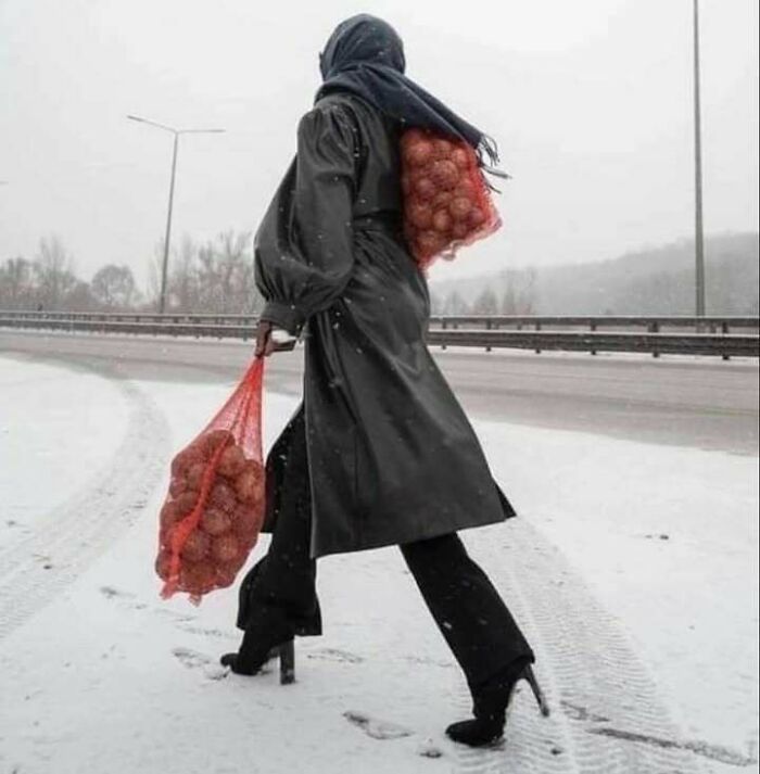 Person walking in snow wearing high heels and a black coat, carrying two large bags of onions, combining trash and culture.