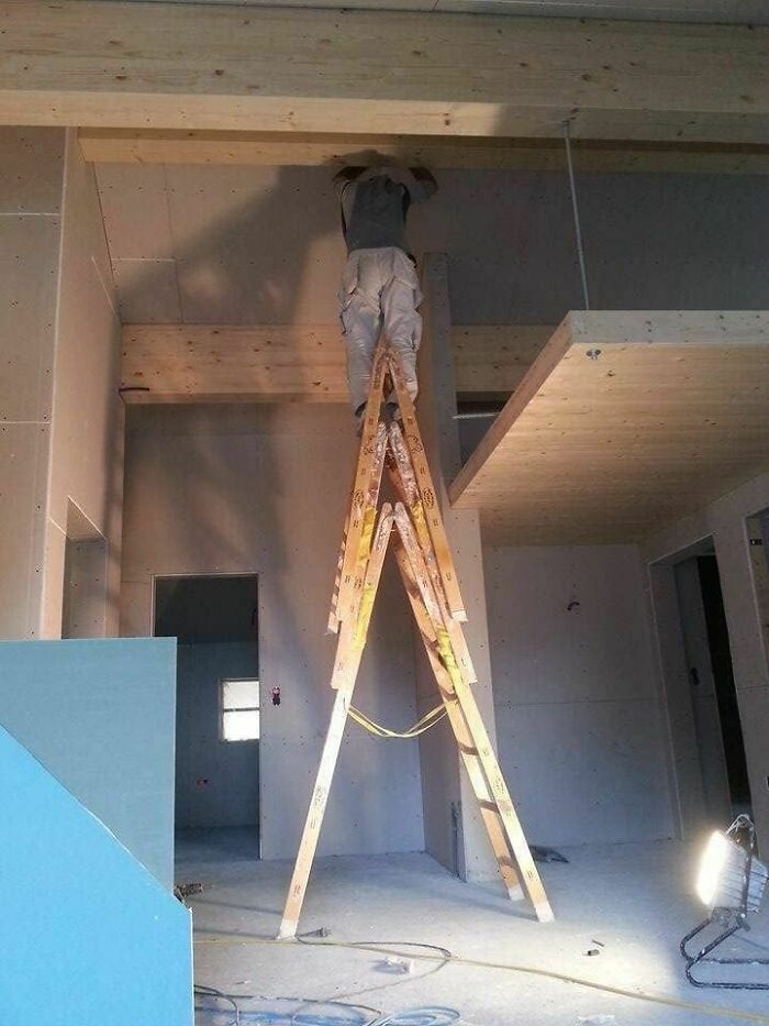 Worker standing unsafely on top of a wooden ladder making a hilariously dumb engineering mistake indoors.