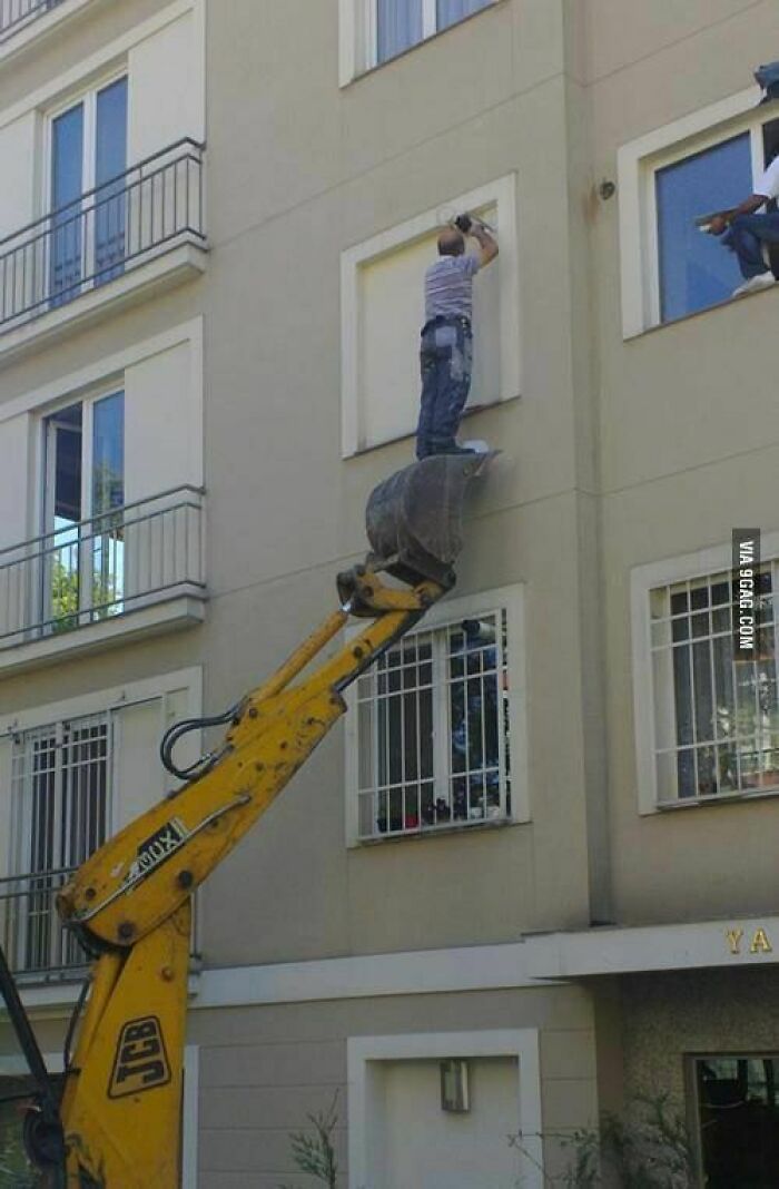 Man standing dangerously on a digger bucket fixing window frame, showing hilariously dumb engineering mistakes in construction work.