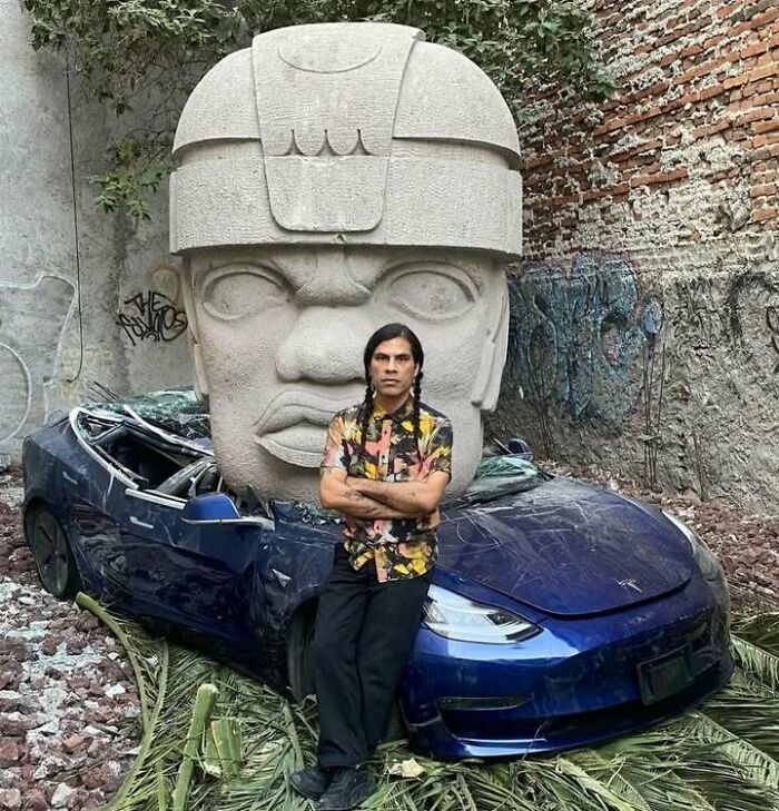 Man in a colorful shirt leaning on a crushed blue car in front of a large stone sculpture combining trash and culture.
