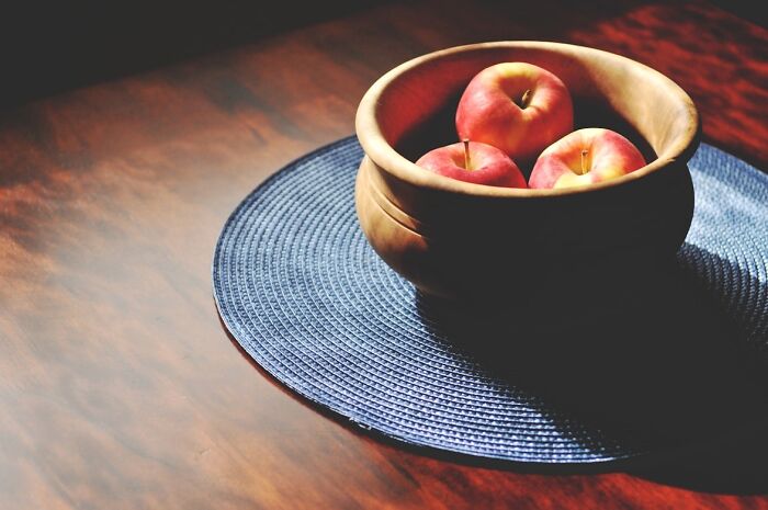 Wooden bowl filled with apples placed on a blue woven mat on a polished wooden table surface.