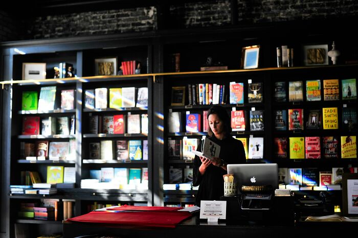 A woman reading a book in a dimly lit bookstore filled with shelves, highlighting red flag romanticized behavior. - 16