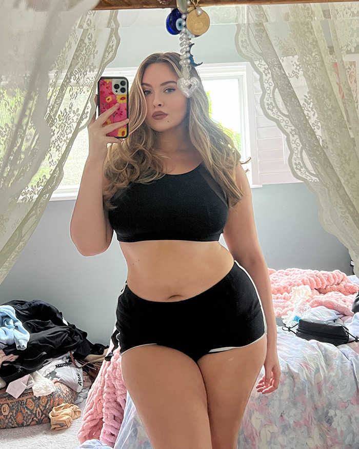 Plus-size model in black crop top and shorts taking a mirror selfie, confidently addressing fatphobic critics.