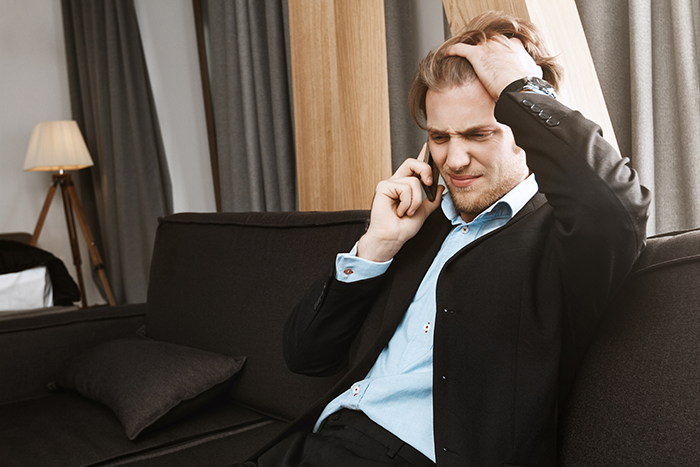 Gay man in suit sitting on couch, looking distressed during phone call about wedding invitation misunderstanding.
