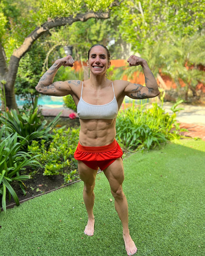 Powerlifter Stefi Cohen flexing muscles outdoors in white top and red shorts, showcasing strength and fitness.