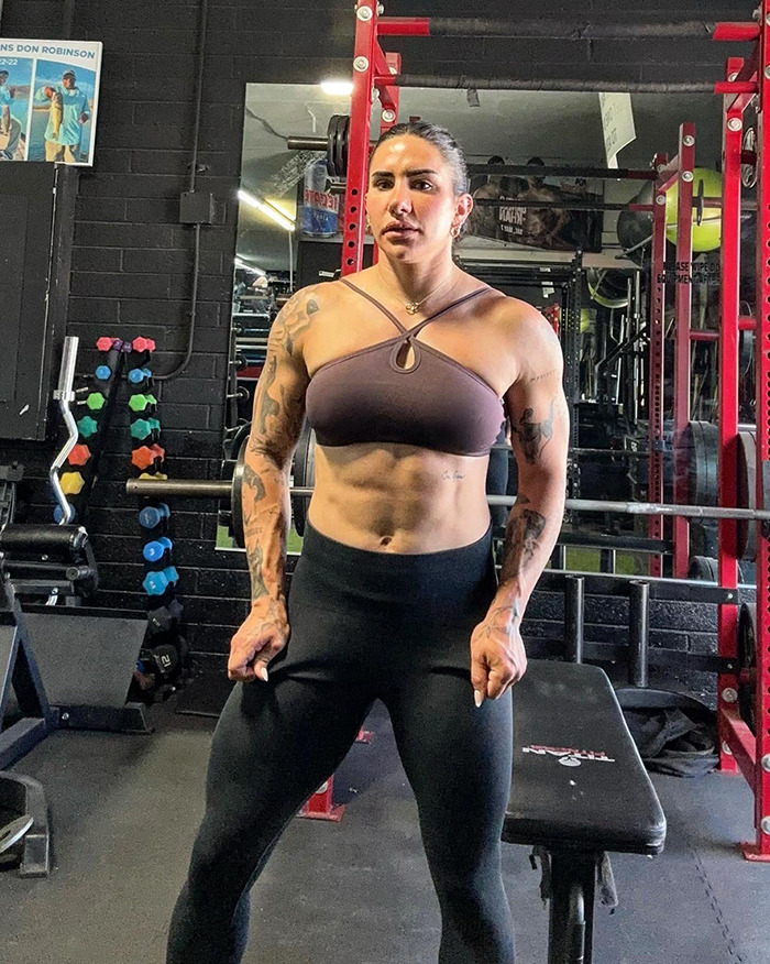 Powerlifter Stefi Cohen posing in gym, showing muscular physique with tattoos near barbells and weights.