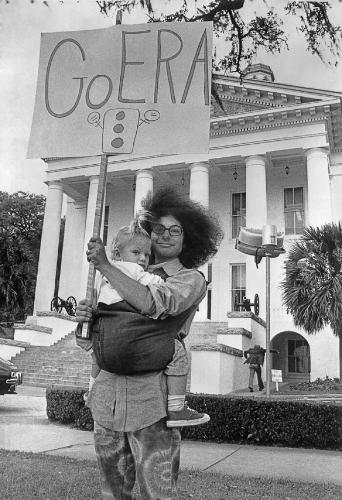 Woman in Florida holding child and protest sign outside a government building, representing women breaking barriers in the 1970s.