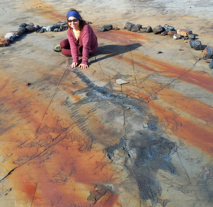 Woman in purple jacket examining a sea reptile fossil over 100 million years old on a rust-colored rock surface outdoors