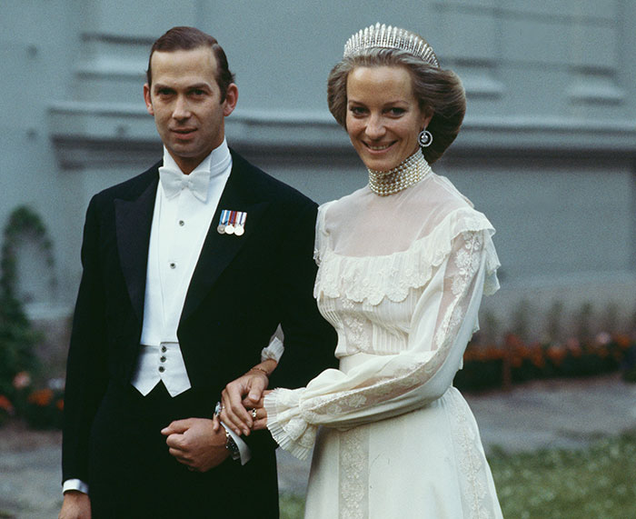 Prince in formal attire holding hands with a smiling royal woman in a white vintage wedding dress and tiara outdoors.