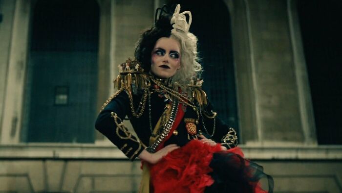 Character Cruella in a dramatic pose wearing a black and red outfit in a dark urban setting for Rate Disney Movies poll