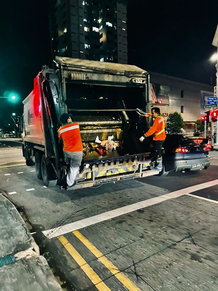Two sanitation workers in orange uniforms collecting trash at night on a city street for make 6 figures jobs.