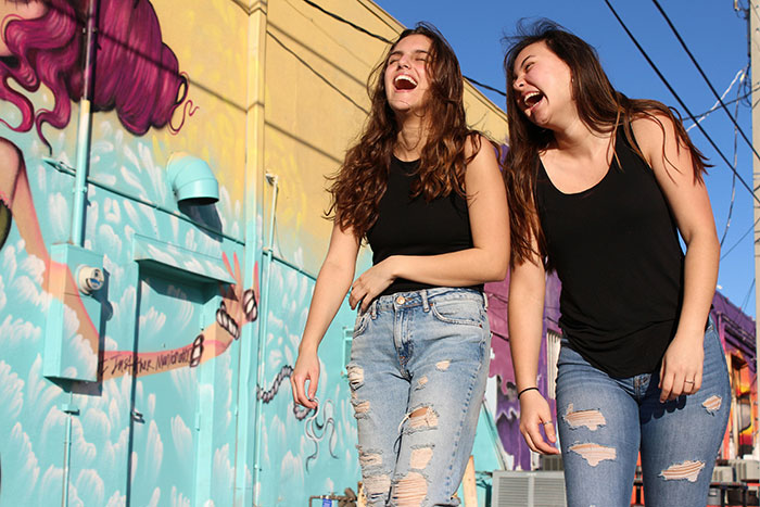 Two young women laughing and walking by a colorful mural, sharing moments with anonymous confessions theme.