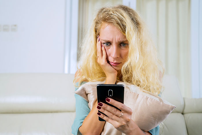 Woman with blonde hair looking shocked and anxious while reading anonymous confessions on her phone at home.