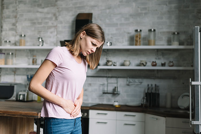 Young woman in a kitchen holding her stomach in discomfort, illustrating anonymous confessions that could ruin lives.