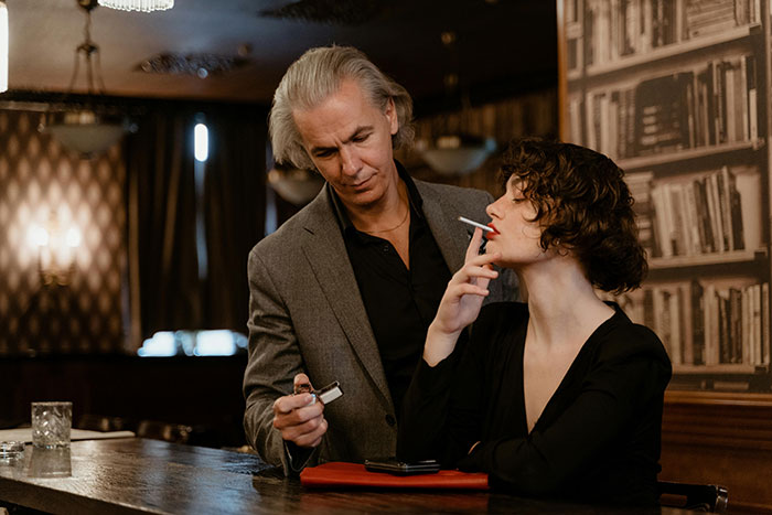 Two people in a dim bar setting, the woman smoking a cigarette while the man holds a lighter, evoking anonymous confessions.