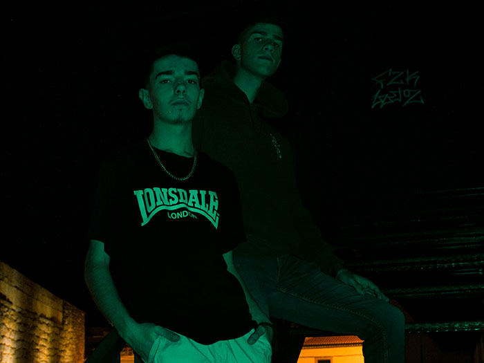 Two young men in a dark, urban setting with graffiti, reflecting signs to look out for unsafe neighborhoods.
