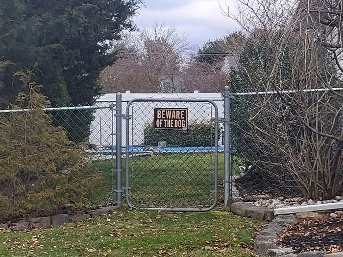 Chain link gate with beware of the dog sign, surrounded by trees and bushes in a neighborhood setting.