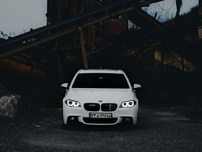 White BMW car with headlights on parked in a dark, abandoned area showing signs of unsafe neighborhoods.