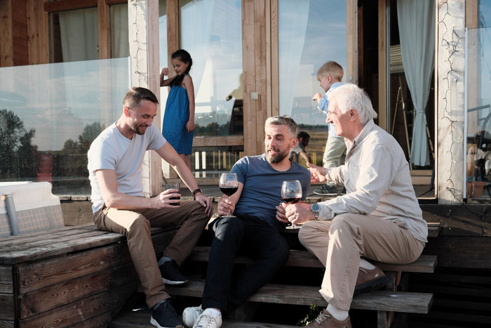 Three men enjoying wine on wooden steps while two children play nearby outside a house on a sunny day.