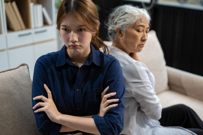 Teen refuses unpaid babysitting and family trip, looking upset while sitting back-to-back with an older woman at home.