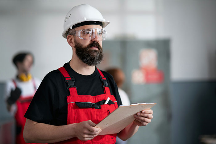 Bearded employee in safety gear holding clipboard at worksite, reflecting on wage reduction and decision to leave company. - 1