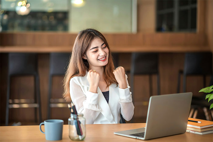 Young woman with PhD celebrating success at laptop in modern indoor workspace with coffee cup and notebooks on table - 13