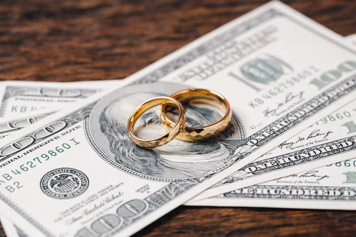 Two gold wedding rings placed on top of hundred-dollar bills representing groom discount and money issues in wedding events. - 10