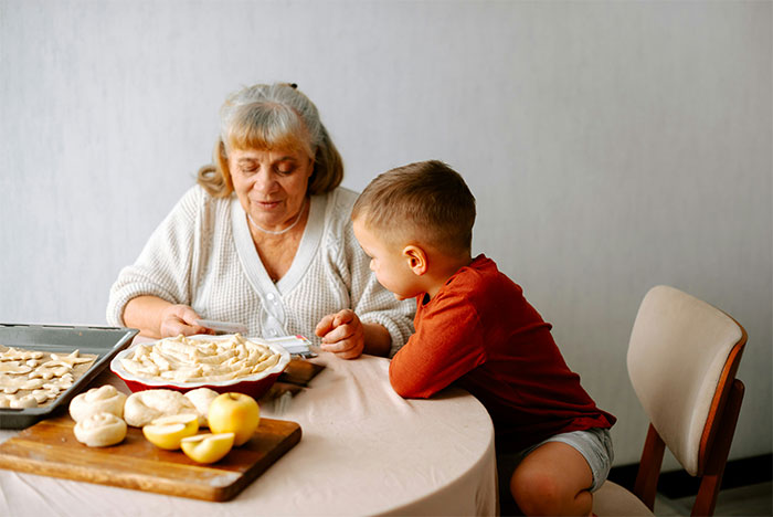 Elderly woman babysitting toddler while baking at a table, highlighting babysitting and toddler care themes.