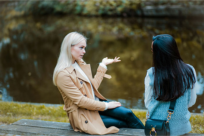 Two women sitting outdoors near water, one angrily explaining while the other listens, highlighting relationship conflict. - 57