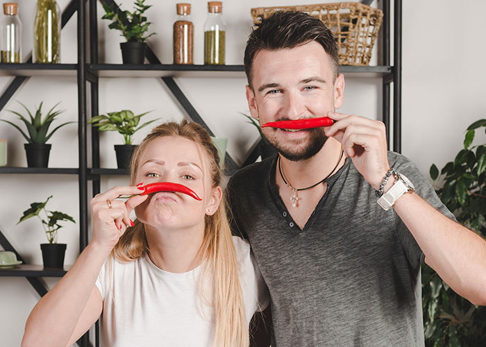 Young couple playfully holding red chili peppers as mustaches in a kitchen setting illustrating seemingly harmless things that are dangerous.