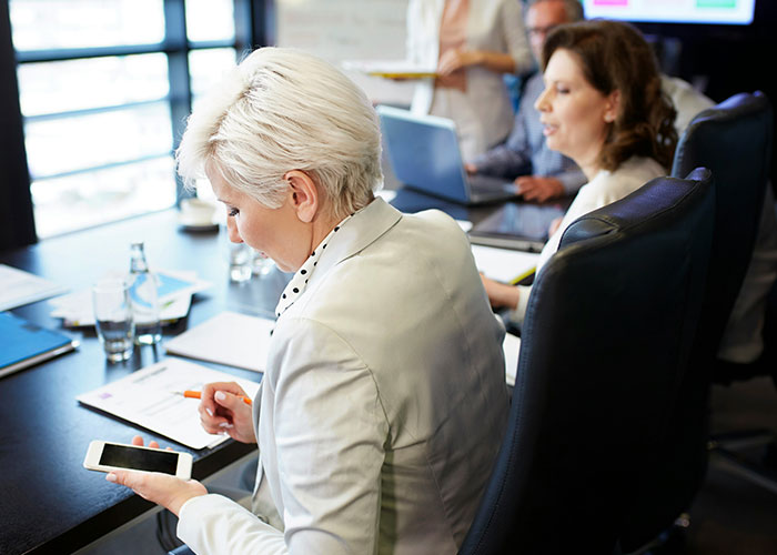 Boss struggling with technology trying to use smartphone in a meeting room with coworkers around a conference table.