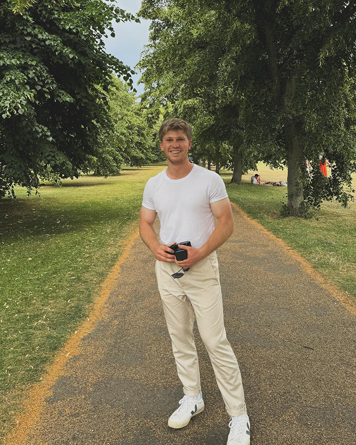Robert Irwin smiling on a park pathway wearing a white shirt, light pants, holding sunglasses and a camera outdoors.