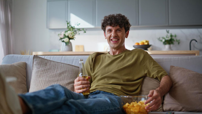 Young man relaxing on couch with beer and chips while watching Jurassic Park, a woman nearby expressing disbelief in dinosaurs.