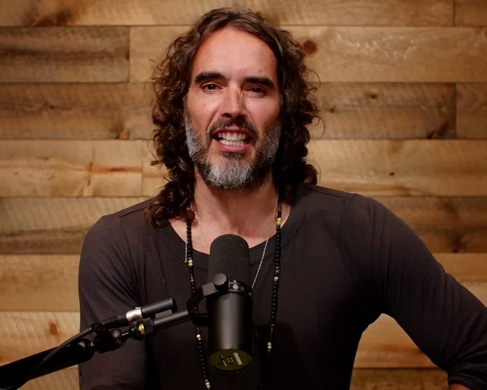 Man with long curly hair and beard speaking into a microphone in front of a wooden wall during a podcast session Man with long curly hair and beard speaking into a microphone in front of a wooden wall during a podcast session