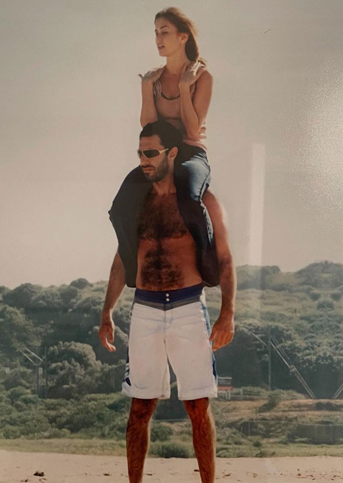 Sacha Baron Cohen shirtless and ripped on beach carrying a woman on his shoulders during a sunny day outdoors.