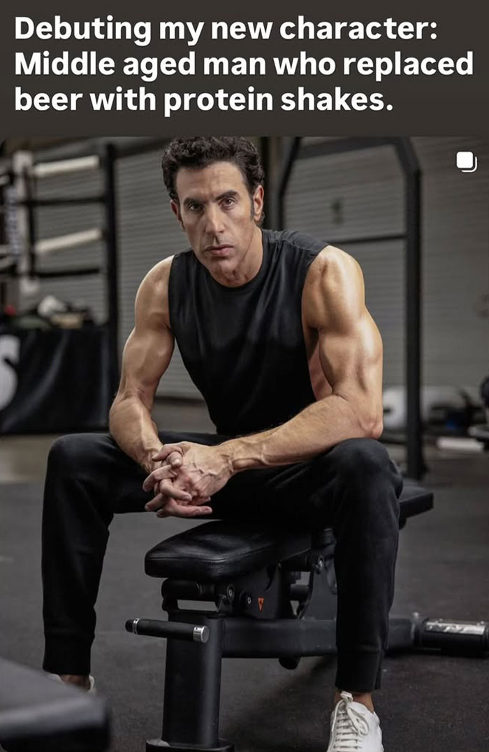 Sacha Baron Cohen showing ripped transformation with muscular arms seated in gym, debuting new character fitness look.