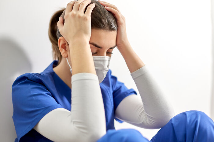 Healthcare worker in blue scrubs and mask holding head in distress, reflecting the challenges of dealing with dead bodies.