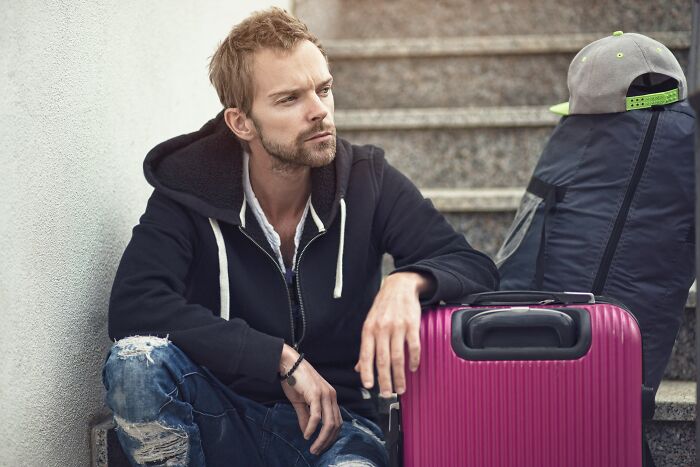 Young man with ripped jeans sitting by stairs next to pink suitcase and backpack, representing student life and outrageous student names.