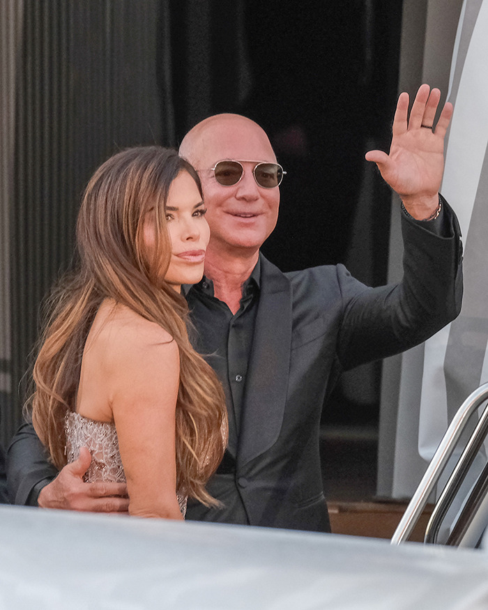 A man in sunglasses waves while standing next to a woman with long hair in an elegant dress, noted for awkward reaction viral.