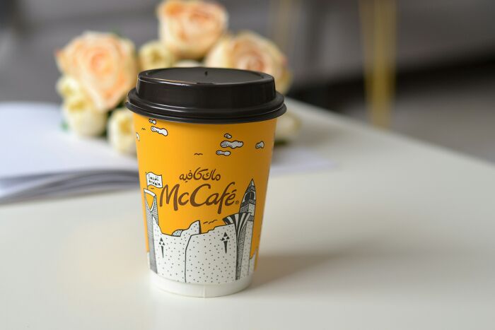 Yellow McCafe coffee cup with a black lid on a white table with blurred roses in the background.
