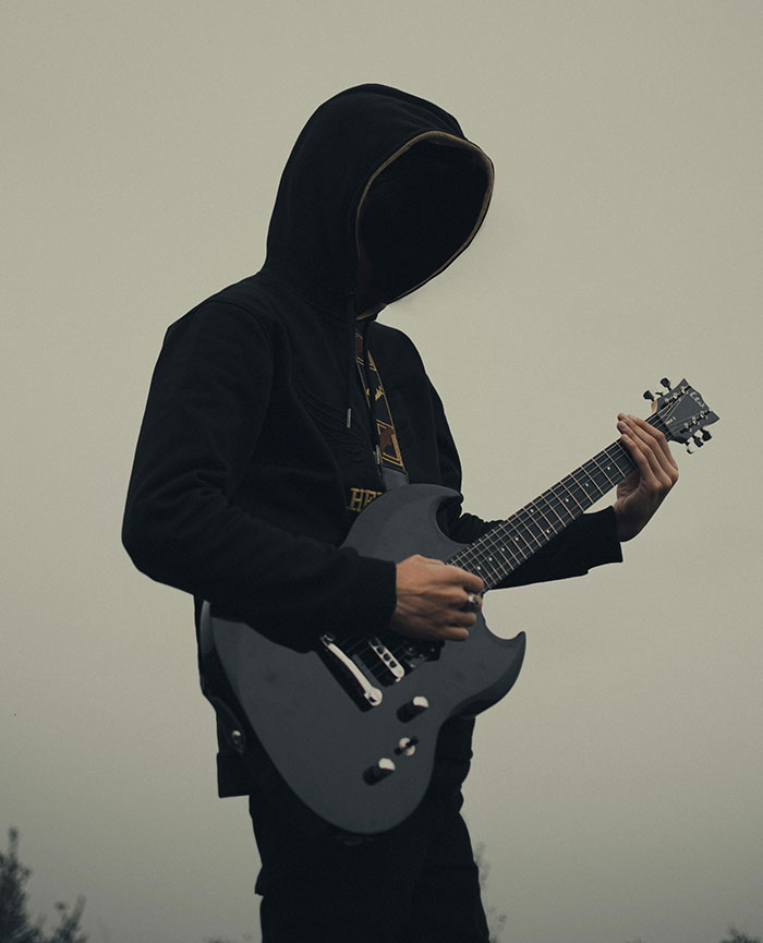Person wearing a black hooded jacket playing an electric guitar outdoors, evoking mysterious neighbor tales in the woods setting.