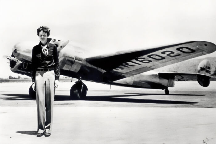 Amelia Earhart standing beside her Lockheed Electra plane in a vintage black and white photograph at an airfield.