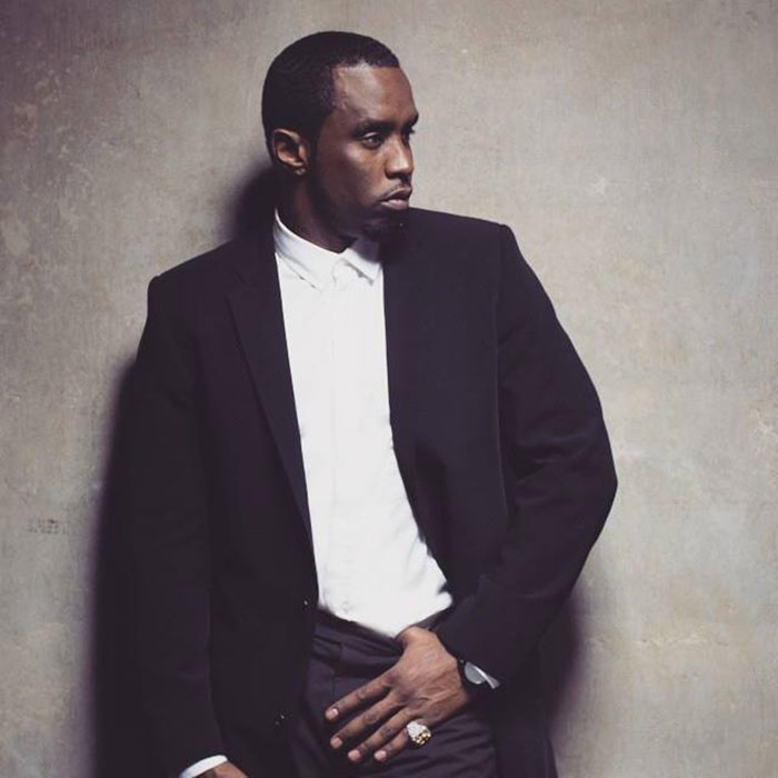 Diddy wearing a black suit and white shirt posing against a textured wall, related to Diddy's trial and jury note incident.