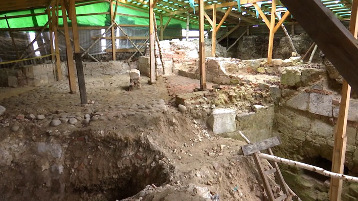 Archaeological excavation revealing a secret chamber beneath castle ruins protected by wooden scaffolding and green tarps.