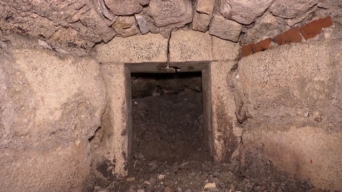 Stone walls and archway leading to a dark secret chamber beneath an ancient castle, partially filled with dirt and debris.