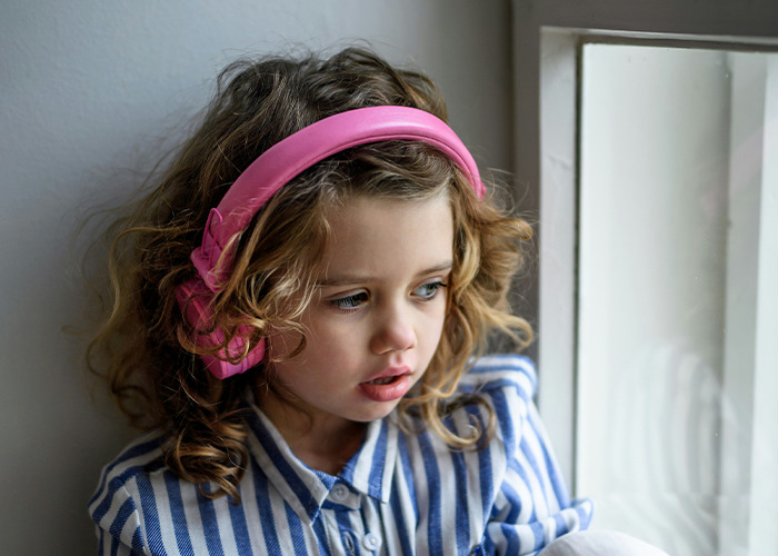 Young girl with curly hair wearing pink headphones and a striped shirt, looking thoughtfully out of a window, secrets hidden. - 6