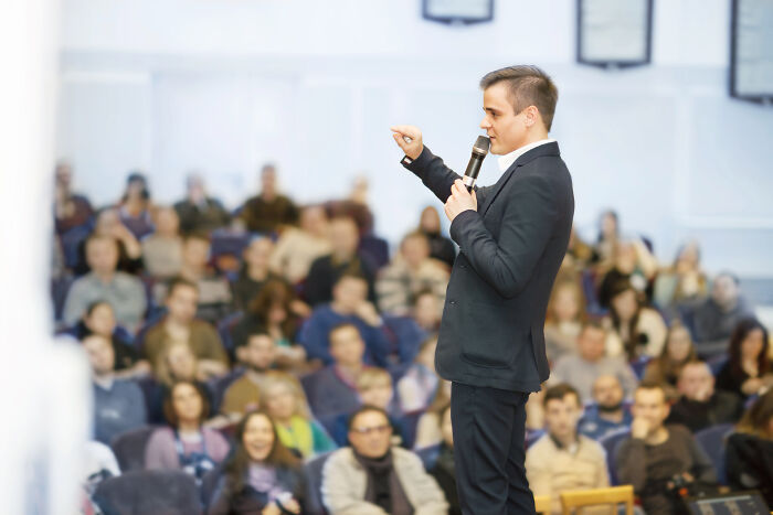Man in a black suit speaking with a microphone to an attentive audience about life-changing sentences.
