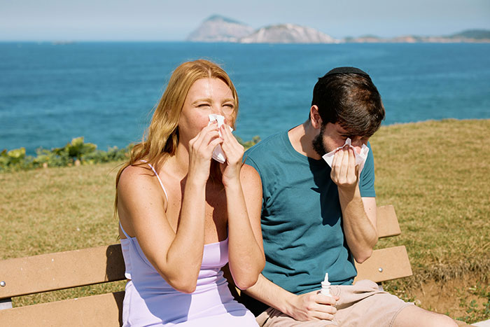 A man and woman sitting outdoors by the sea, both sneezing and using tissues, indicating symptoms of serious medical conditions.