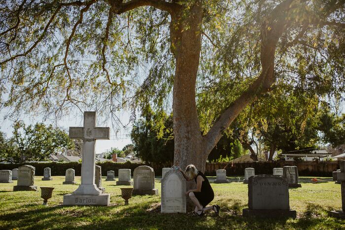 Woman mourning by a grave in a sunlit cemetery with large trees, reflecting signs you grew up sheltered emotions.